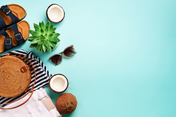 Female birkenstock sandals, jeans, striped t-shirt, rattan bag, coconut and sunglasses on blue background with copy space. Top view. Summer fashion, capsule wardrobe concept. Creative flat lay